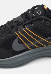 Chaussure de sport noire avec une tige en tissu texturé, des accents de rayures orange et une semelle grise rembourrée, dotée d'œillets circulaires et de lacets.