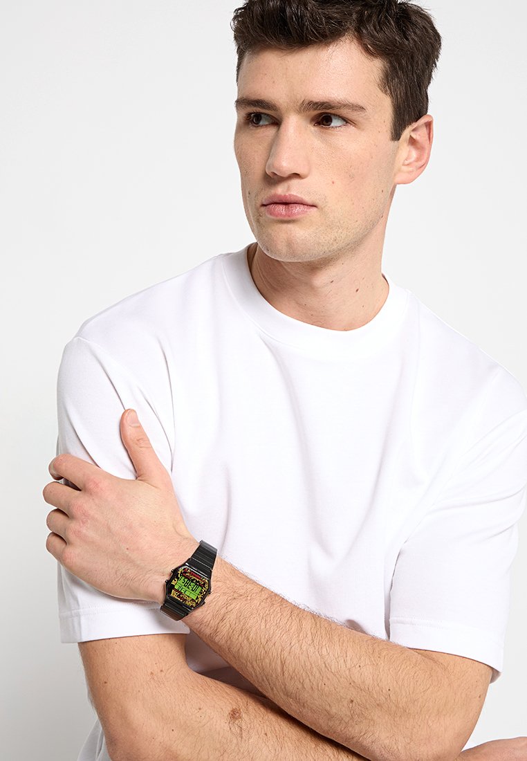 Reloj digital con correa negra, esfera cuadrada en dorado y verde, y tipografía en negrita. Llevo puesto en la muñeca sobre una camiseta blanca lisa.