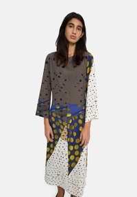 Młoda kobieta z długimi ciemnymi włosami, ubrana w długą, luźną sukienkę w abstrakcyjne wzory w różne kropki w kolorach brązowym, niebieskim, białym i złotym.