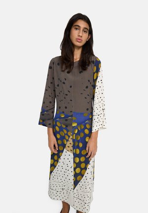 Młoda kobieta z długimi ciemnymi włosami, ubrana w długą, luźną sukienkę w abstrakcyjne wzory w różne kropki w kolorach brązowym, niebieskim, białym i złotym.