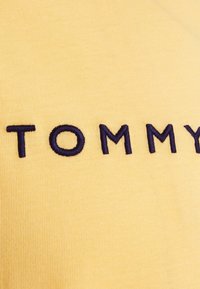 Tecido de algodão amarelo com bordado do texto "TOMMY" em azul marinho. A textura é suave, com um acabamento canelado.