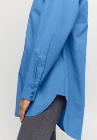 Camicia blu con colletto a punta, orlo arrotondato, maniche lunghe e una texture liscia e leggera. Presenta bottoni ai polsini per un tocco di dettaglio.