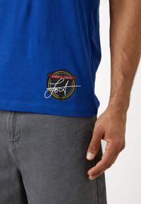 Chemise bleue avec un logo circulaire noir, jaune et rouge près de l'ourlet portant le nom "Nikola Jokić" et une signature blanche stylisée.
