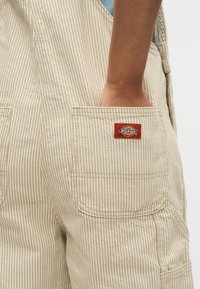 Main dans la poche arrière d'une salopette Dickies beige à rayures blanches avec un patch logo rouge visible.