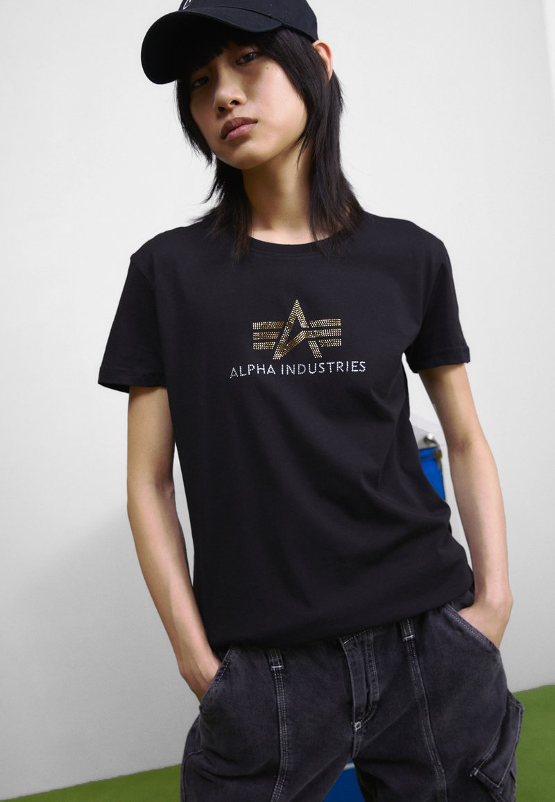 T-shirt noir avec un logo métallique doré "ALPHA INDUSTRIES" et des motifs géométriques. Le modèle porte un pantalon cargo sombre et une casquette noire.