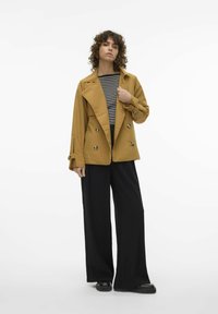 Sennepsgul dobbeltknappet blazer med store knapper, over en sort og hvid stribet skjorte. Kombineret med brede sorte bukser og sorte sko.