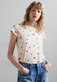 T-Shirt krem bo kratkim rukavima s višebojnim uzorkom gljiva. Izrađen od mekog materijala s klasičnim krojem, u kombinaciji s plavim trapericama.