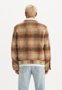Chaqueta de cuadros marrón y beige con un cuello de borrego, que presenta un ajuste estructurado y cierres de botón. Llevada sobre jeans de color azul claro.