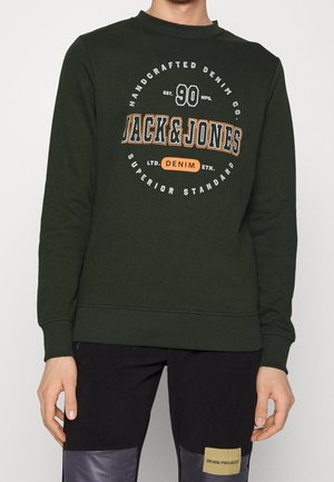 Donkergroene trui met circulaire "Jack & Jones" denim branding in witte en oranje tekst op de borst, gedragen met zwarte broek.