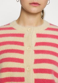 Hřejivý kardigan se širokými horizontálními pruhy v růžové a krémové barvě, s kulatým výstřihem a třemi velkými knoflíky vpředu.