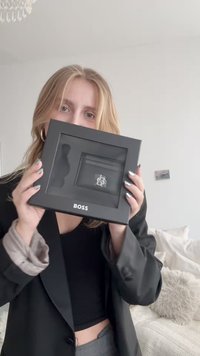 Czarne prostokątne pudełko z przezroczystą pokrywą, zawierające małą czarną kopertówkę i srebrny element ozdobny. Widoczne wyraźnie logo „BOSS”.