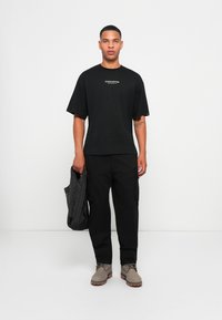 Zwarte oversized t-shirt met de tekst "STUDIO EDITION", gecombineerd met zwarte cargobroek en lichtgrijze veterschoenen. Houdt een zwarte tas vast.