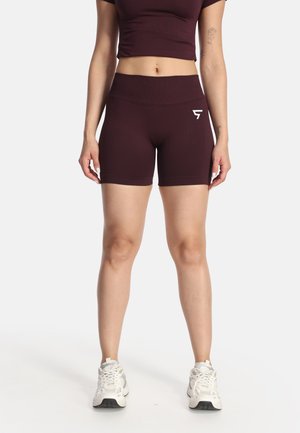 Donna che indossa pantaloncini da biker a vita alta color granata e top corto abbinato, in piedi con scarpe sportive bianche su sfondo bianco.