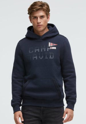 Young man with light brown hair wearing a navy blue hooded sweatshirt with "Camp David" text and two small patches on the chest.