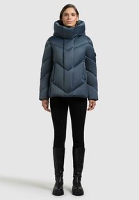 Piumino blu scuro con collo alto, motivo a zig zag, tasche con zip, indossato sopra leggings neri e stivali al ginocchio.