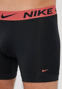 Sorte boxer briefs til mænd lavet af glat stof med et rødt Nike-logo i taljen og en lille pink Nike swoosh. De har en tætsiddende pasform og korte ben.