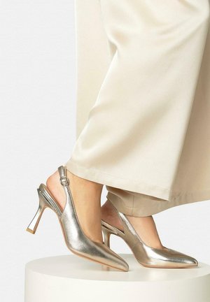 Tacones altos de punta puntiaguda plateados metálicos con tira trasera, usados con pantalones anchos color crema, pies descansando sobre una plataforma redonda blanca.