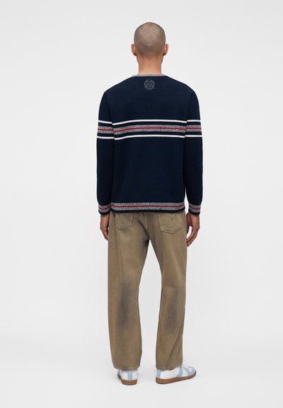 Homme à la tête rasée portant un pull bleu marine avec des rayures horizontales blanches et rouges, un pantalon beige et des sandales blanches, debout de dos.