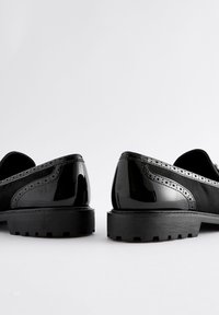 Schwarze Lederschuhe mit glänzendem Finish, niedrigem Absatz, strukturierter Gummisohle und dekorativen perforierten Details entlang der Ränder.