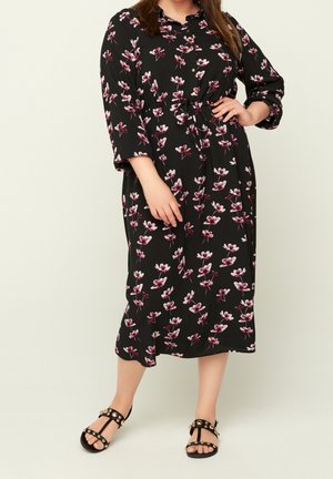 Nainen pukeutuneena mustaan midi-mekkoon, jossa on vaaleanpunaisia ja valkoisia kukkakuvioita. Mekossa on puolipitkät hihat. Hän seisoo käsi lanteilla ja jalassa on mustat niititiet sandaalit.