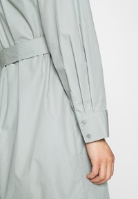 Svetle šedé šaty s páskem a sklady, s knoflíkovým manžetovým rukávem a hladkým, strukturovaným materiálem.