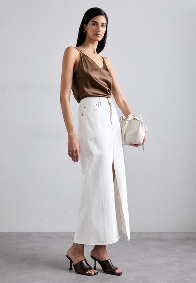 White maxi skirt with a front slit, paired with a brown satin camisole. The model holds a white bag with a bow and wears dark mules.