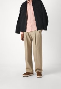 Svart zip-up jacka, ljusrosa skjorta med knappar, beige plisserade byxor, bruna sneakers med vita sulor, mot en neutral bakgrund.