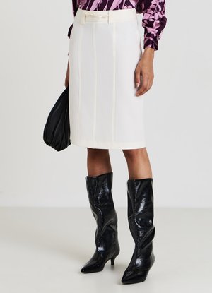 Weißer Bleistiftrock mit vertikalen Nähten und tailliertem Bund, kombiniert mit glänzenden schwarzen Kniefallstiefeln, gehalten mit einer schwarzen Tasche.