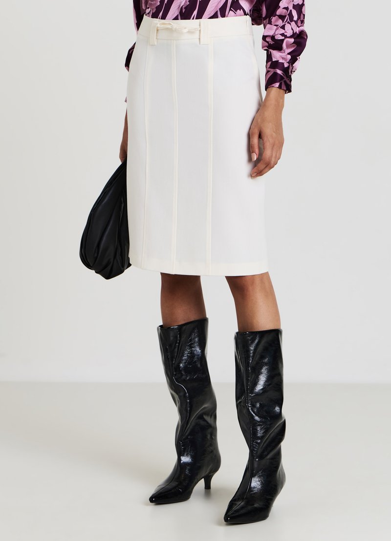 Weißer Bleistiftrock mit vertikalen Nähten und tailliertem Bund, kombiniert mit glänzenden schwarzen Kniefallstiefeln, gehalten mit einer schwarzen Tasche.