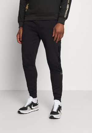 Mann trägt schwarze Jogginghose, schwarzen Pullover mit goldenen Schriftzügen auf den Ärmeln, weiße Socken und schwarz-weiße Nike-Sneaker.