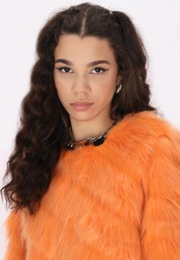 Orange faux furtröja med hög krage, som har en svart kedjedetalj och textil med volym. Lockigt hår som ramar in ansiktet.