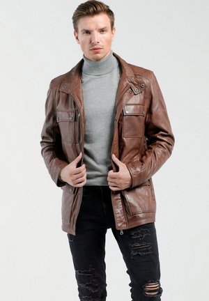 Braune Lederjacke mit Reißverschluss, zwei Brusttaschen und hohem Kragen. Getragen über einem grauen Rollkragenpullover und schwarzen, zerrissenen Jeans.