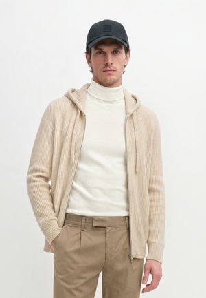 Uomo che indossa un berretto nero, una felpa beige in maglia con zip, un dolcevita bianco e pantaloni beige, in piedi su uno sfondo bianco semplice.