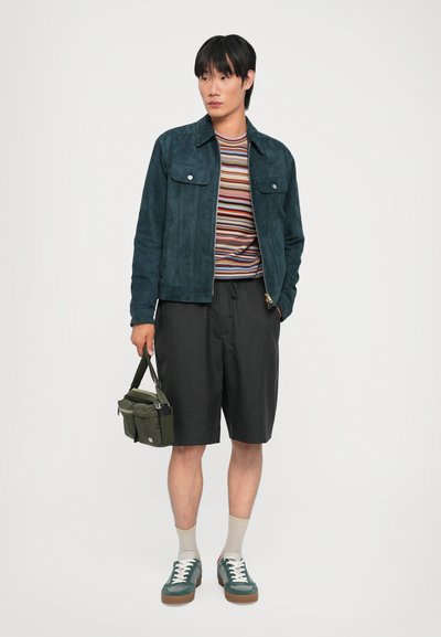 Veste en suède bleu canard, t-shirt rayé multicolore, shorts foncés, baskets vertes avec lacets blancs, tenant un sac vert avec plusieurs compartiments.