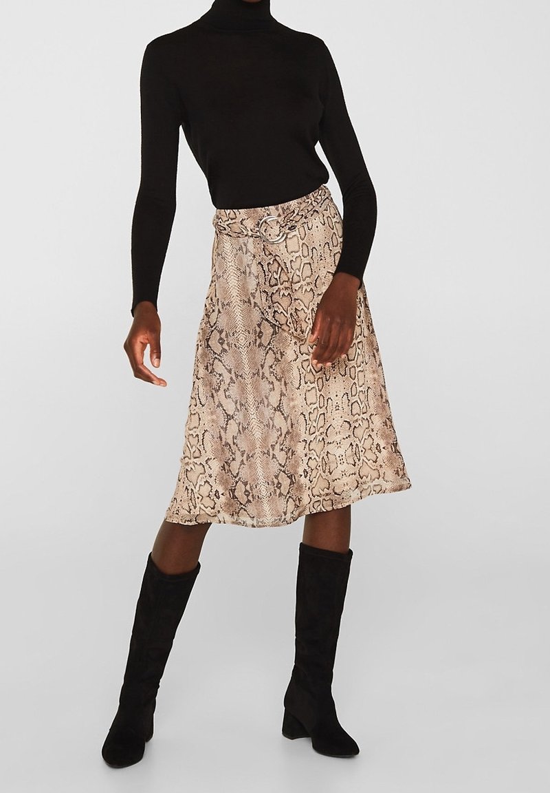 Femme portant un haut noir à manches longues, une jupe midi beige à imprimé serpent avec ceinture, et des bottes noires montant jusqu'au genou, sur un fond uni.