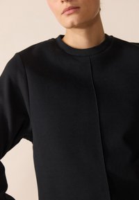 Svart genser laget av teksturert stoff, med rund hals og en søm som går nedover midten for et rent og minimalistisk design.