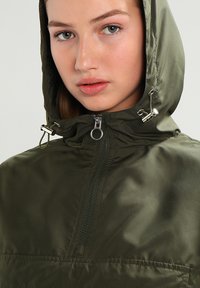 Ung kvinne med blågrønne øyne som har på seg en olivengrønn hettejakke med glidelås og metalliske snorer.