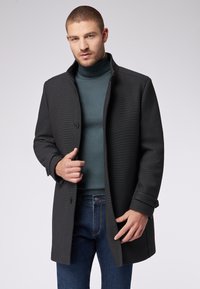 Dunkelgrauer, strukturierter Mantel mit hohem Kragen, einzelnem Knopfverschluss und hochgekrempelten Ärmeln, getragen über einem türkisfarbenen Rollkragenpullover und blauen Jeans.