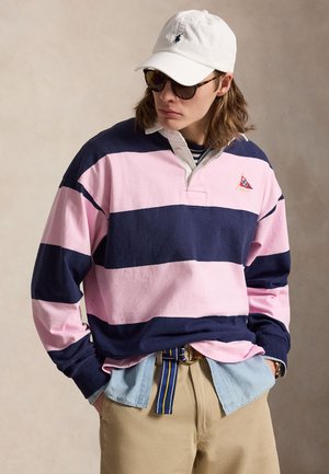 Isik, kes kannab valget nokamütsi, tumedaid päikseprille, roosa ja mereväe triibulist ragbimängusärki, mis on kihiliselt kerge teksasärgiga, beežide pükste ja triibulise vööga.