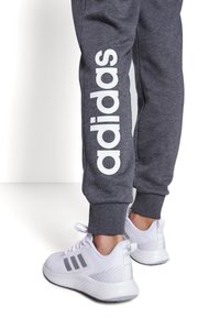 Un pantalon de survêtement de sport gris présente un grand logo "adidas" blanc sur la jambe gauche, associé à des baskets blanches mettant en valeur des rayures gris accentuées.