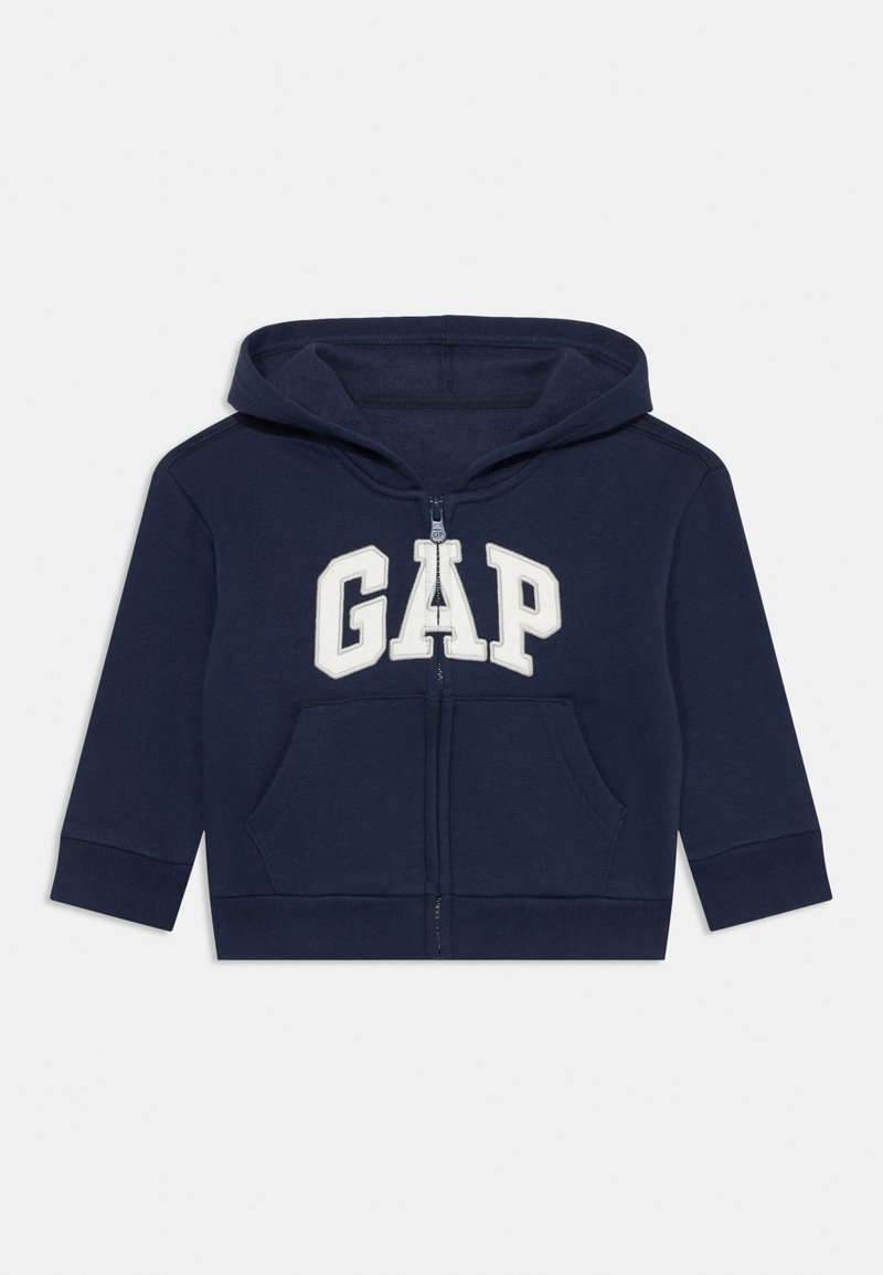 Tmavě modrá mikina na zip vyrobená z bavlněné látky, s velkým bílým logem "GAP" na přední straně a dvěma předními kapsami.