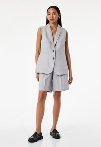 Chaleco gris ajustado con un escote en V profundo y dos bolsillos frontales, combinado con pantalones cortos a la rodilla a juego y mocasines negros. Textura suave.