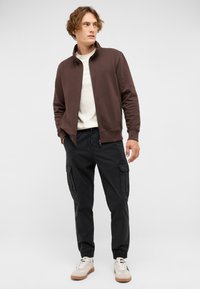 Young man looking sideways wearing a brown zip-up jacket, white shirt, black cargo pants, and light-colored sneakers on a plain background.