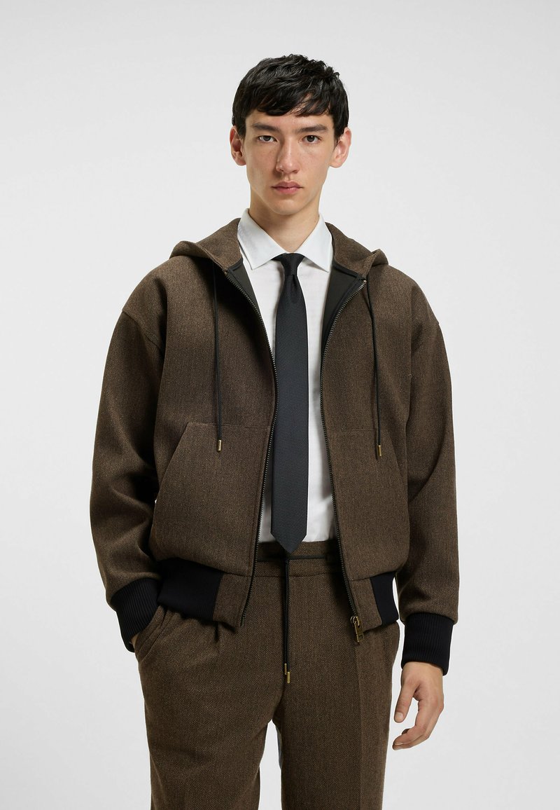 Bruine, gestructureerde hooded jacket met een rits aan de voorkant, twee zakken en zwarte accenten; gedragen over een wit shirt en stropdas, gecombineerd met bijpassende broek.