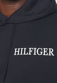 Sudadera negra de algodón, con un logo blanco de “HILFIGER” en el pecho, capucha con cordón y tejido texturizado.