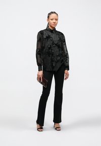Čierna priehľadná blúzka s kvetinovou výšivkou, dlhými rukávmi a vysokým golierom, kombinovaná s čiernymi priliehavými nohavicami a čiernymi sandálmi na podpätku.