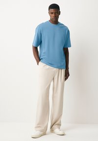 Texturált anyagból készült kék rövid ujjú póló, világos bézs, laza szabású nadrággal és fehér tornacipővel párosítva. Egyszerű kialakítás, minta nélkül.