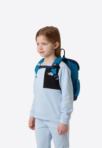Zils mugursoma ar melniem akcentiem, regulējamām siksnām un uzglabāšanas kabatām. Valdzēta bērna gaiši zilos krekliņā un atbilstošās biksēs.