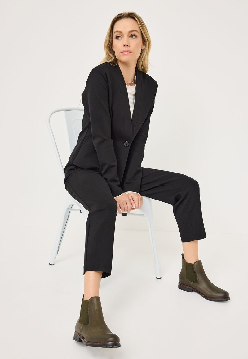 Blazer noir ajusté sur un haut rayé, associé à des bottines en cuir vert foncé avec des panneaux élastiques sur les côtés, assise sur une chaise en métal.