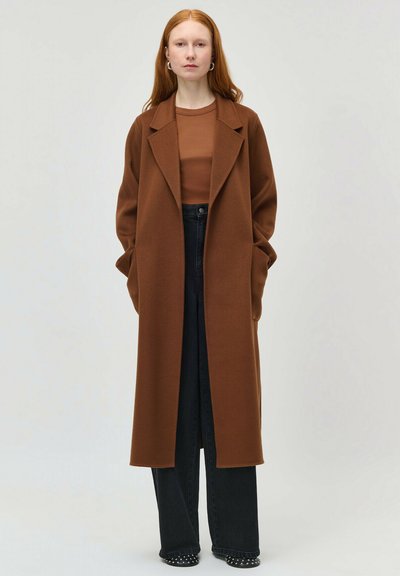 Femeie cu păr roșu purtând o haină lungă maro, bluză maro, pantaloni negri cu croială largă și pantofi negri cu ținte, stând pe un fundal simplu.